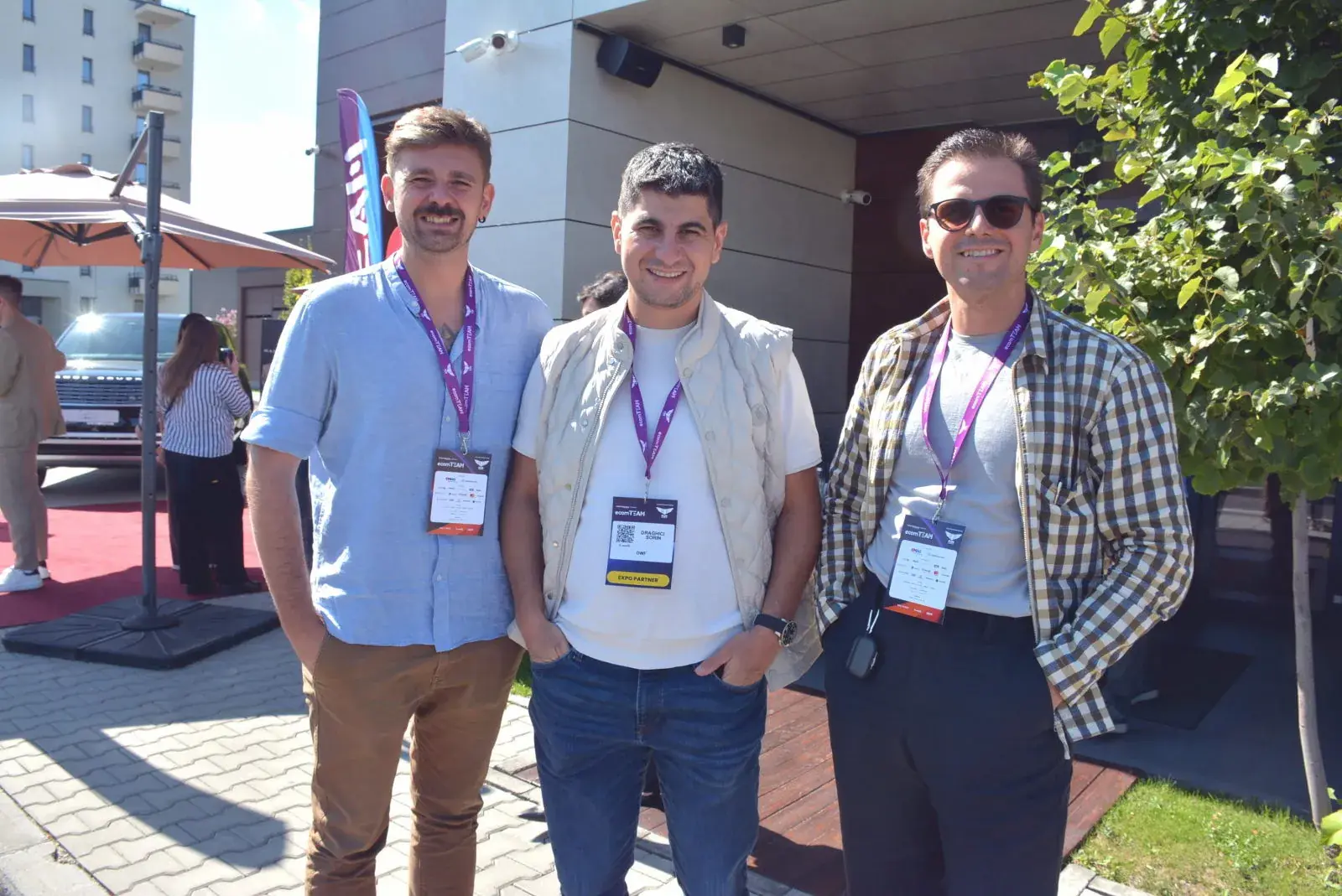 Trei participanți cu ecusoane ecomTEAM zâmbesc la o discuție în aer liber, în pauză.