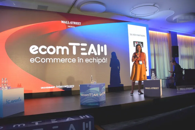 Scena ecomTEAM, cu mesajul „eCommerce în echipă” pe ecranul mare și o speakeriță la microfon.